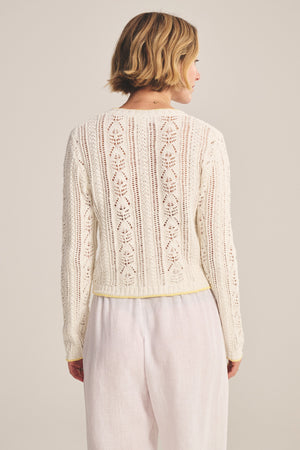 A person with short blonde hair is seen from the back, wearing Velvet by Graham & Spencer's DAISY CARDIGAN, a long-sleeved white knit with pointelle detailing, paired with white pants.