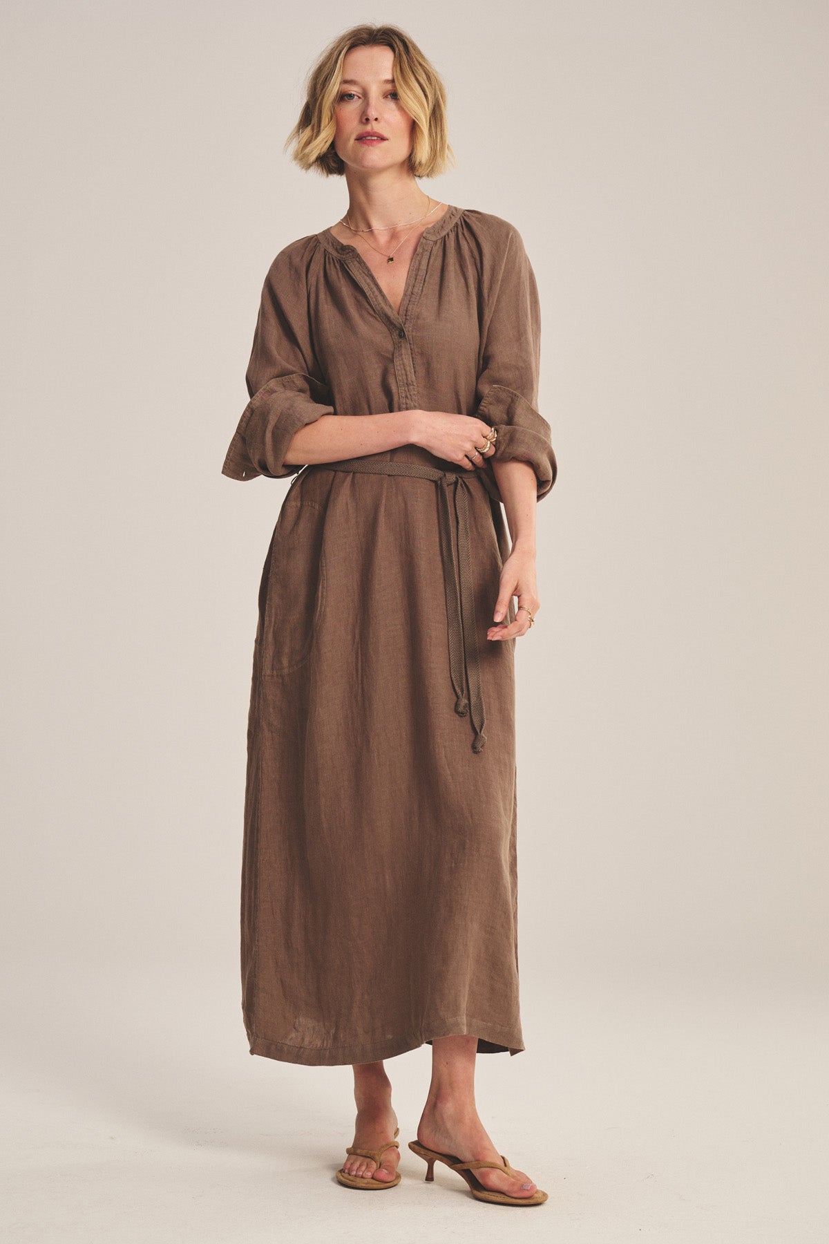 A woman wears the DAKOTA LINEN DRESS by Velvet by Graham & Spencer—a long, breathable linen maxi dress with a detachable belt and long sleeves—paired with tan sandals, against a plain light background.-41081405800641