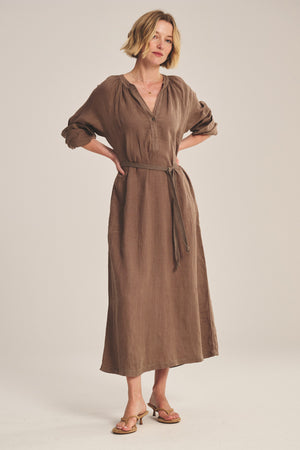 A woman stands with hands on hips, wearing the DAKOTA LINEN DRESS by Velvet by Graham & Spencer—a breathable brown maxi dress with a detachable belt—and sandals, set against a plain background.