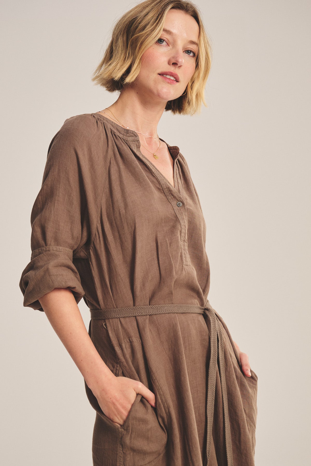 A woman with short blonde hair poses against a plain background wearing the Velvet by Graham & Spencer DAKOTA LINEN DRESS, a loose-fitting, breathable maxi linen dress featuring a detachable belt and pockets.
