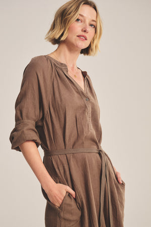 A woman with short blonde hair poses against a plain background wearing the Velvet by Graham & Spencer DAKOTA LINEN DRESS, a loose-fitting, breathable maxi linen dress featuring a detachable belt and pockets.