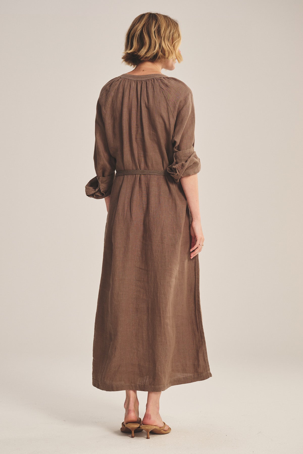A woman faces away, wearing the DAKOTA LINEN DRESS by Velvet by Graham & Spencer— a breathable maxi with detachable belt—paired with tan heeled sandals, set against a plain background.