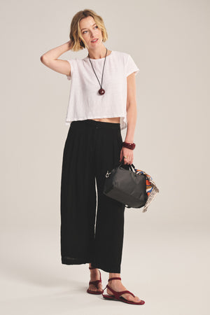A woman stands against a plain background wearing a white cropped t-shirt, Velvet by Graham & Spencer DANIKA LINEN PANT in black, red sandals, and holding a black handbag with a scarf.