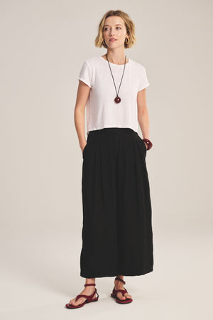A woman stands with hands in her pockets, wearing the DANIKA LINEN PANT by Velvet by Graham & Spencer, a white short-sleeve top, red sandals, and matching jewelry against a plain background.