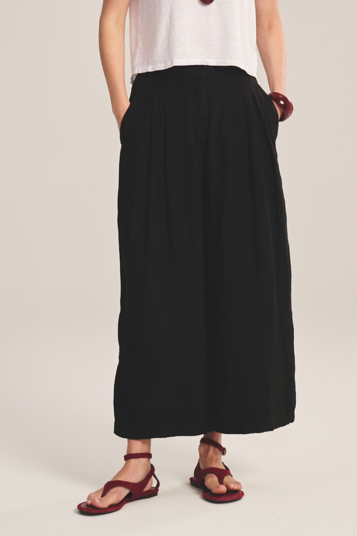 A person wearing a white top and DANIKA LINEN PANT by Velvet by Graham & Spencer, high-waisted and wide-leg in black, paired with red flat sandals, standing with hands in pockets.