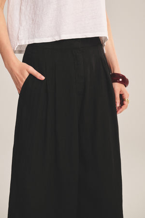 A person wears a white top and the DANIKA LINEN PANT by Velvet by Graham & Spencer—high-waisted, wide-leg black pants with pockets—accessorized with a chunky red bracelet and rings.