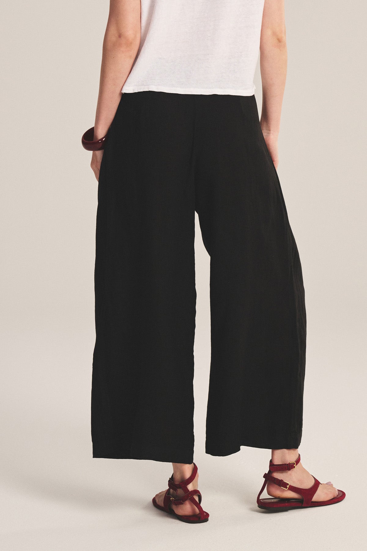 A person shown from the back wearing Velvet by Graham & Spencer's DANIKA LINEN PANT in black, a white short-sleeve shirt, red sandals, and a chunky bracelet.