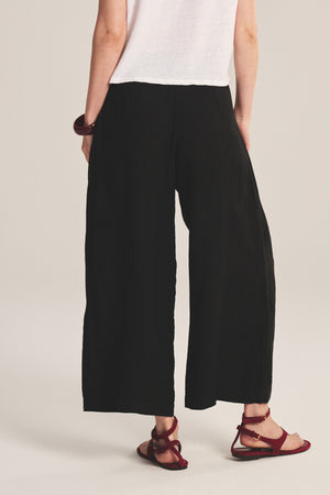 A person shown from the back wearing Velvet by Graham & Spencer's DANIKA LINEN PANT in black, a white short-sleeve shirt, red sandals, and a chunky bracelet.