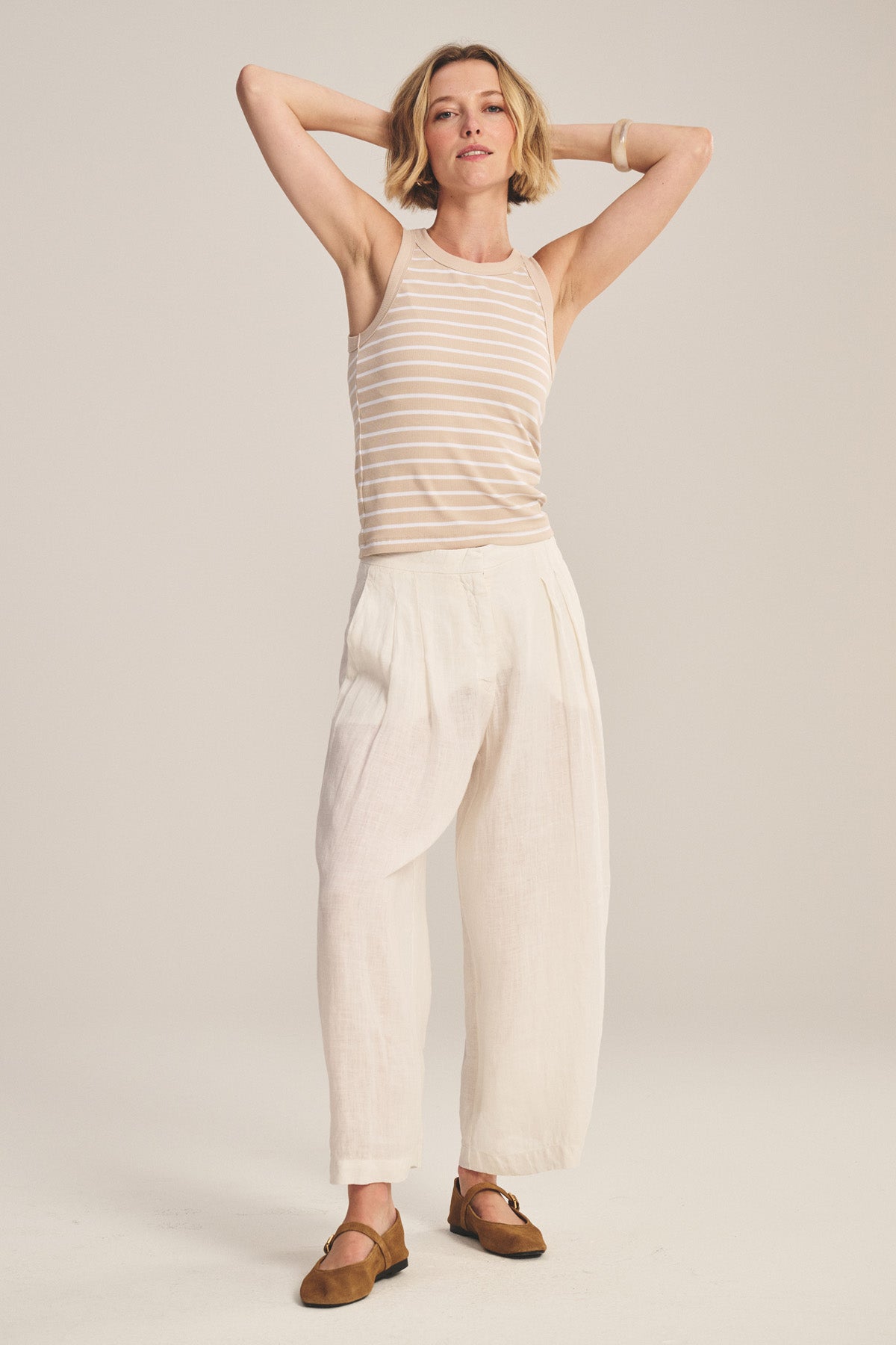 A person stands with arms raised, wearing the DANIKA LINEN PANT by Velvet by Graham & Spencer, paired with a beige and white striped tank top, brown shoes, and a beige bracelet against a plain light background.