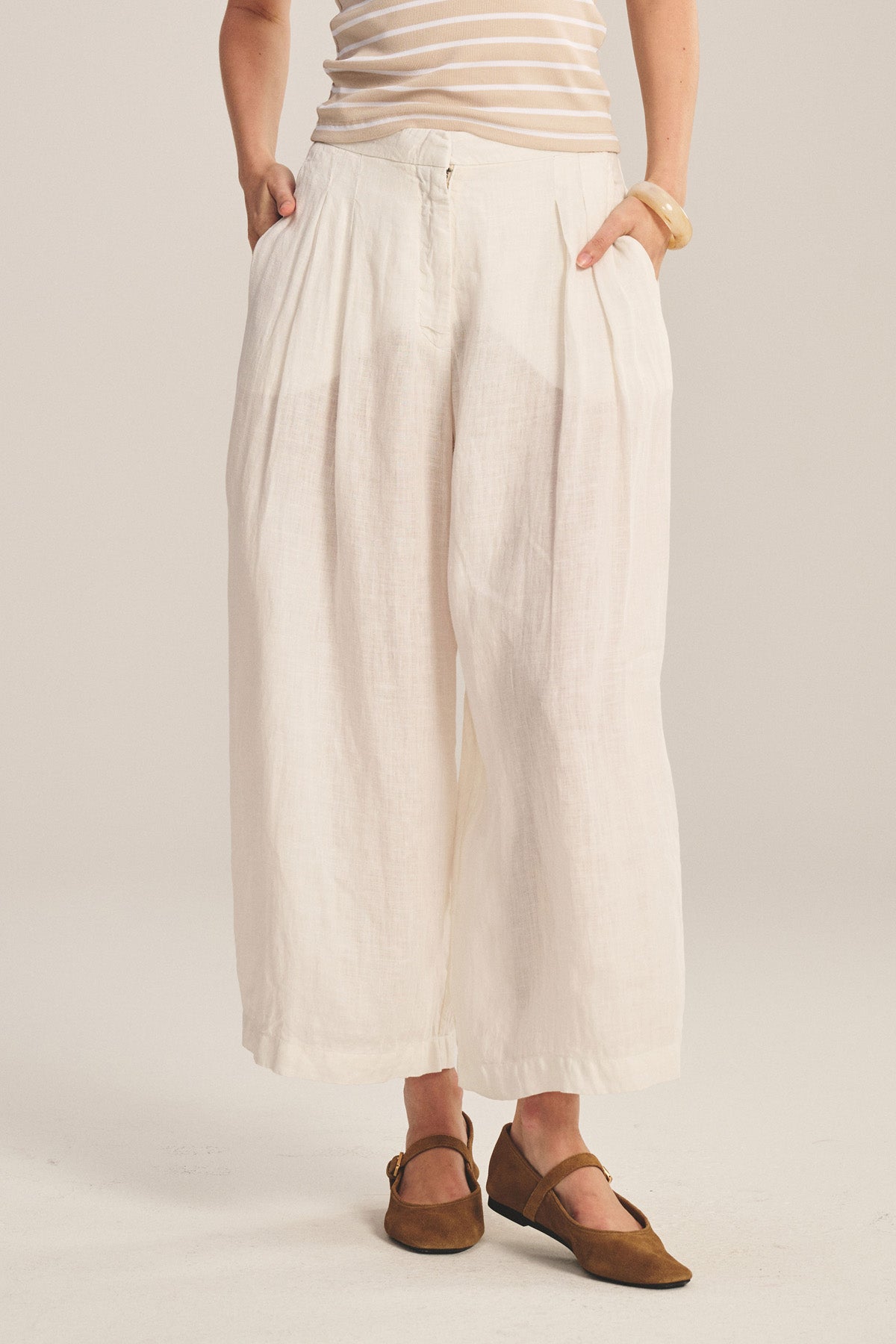 Wearing the DANIKA LINEN PANT by Velvet by Graham & Spencer in white, paired with a beige and white striped top, brown flats, and a light bracelet; hands in pockets against a plain backdrop.