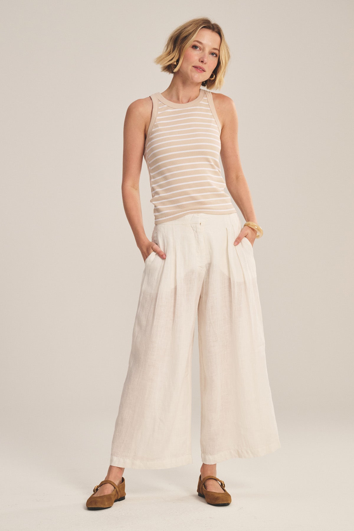 A woman stands with hands in pockets against a plain background, wearing the DANIKA LINEN PANT by Velvet by Graham & Spencer styled with a sleeveless beige and white striped top and brown flat shoes.-41081409732801