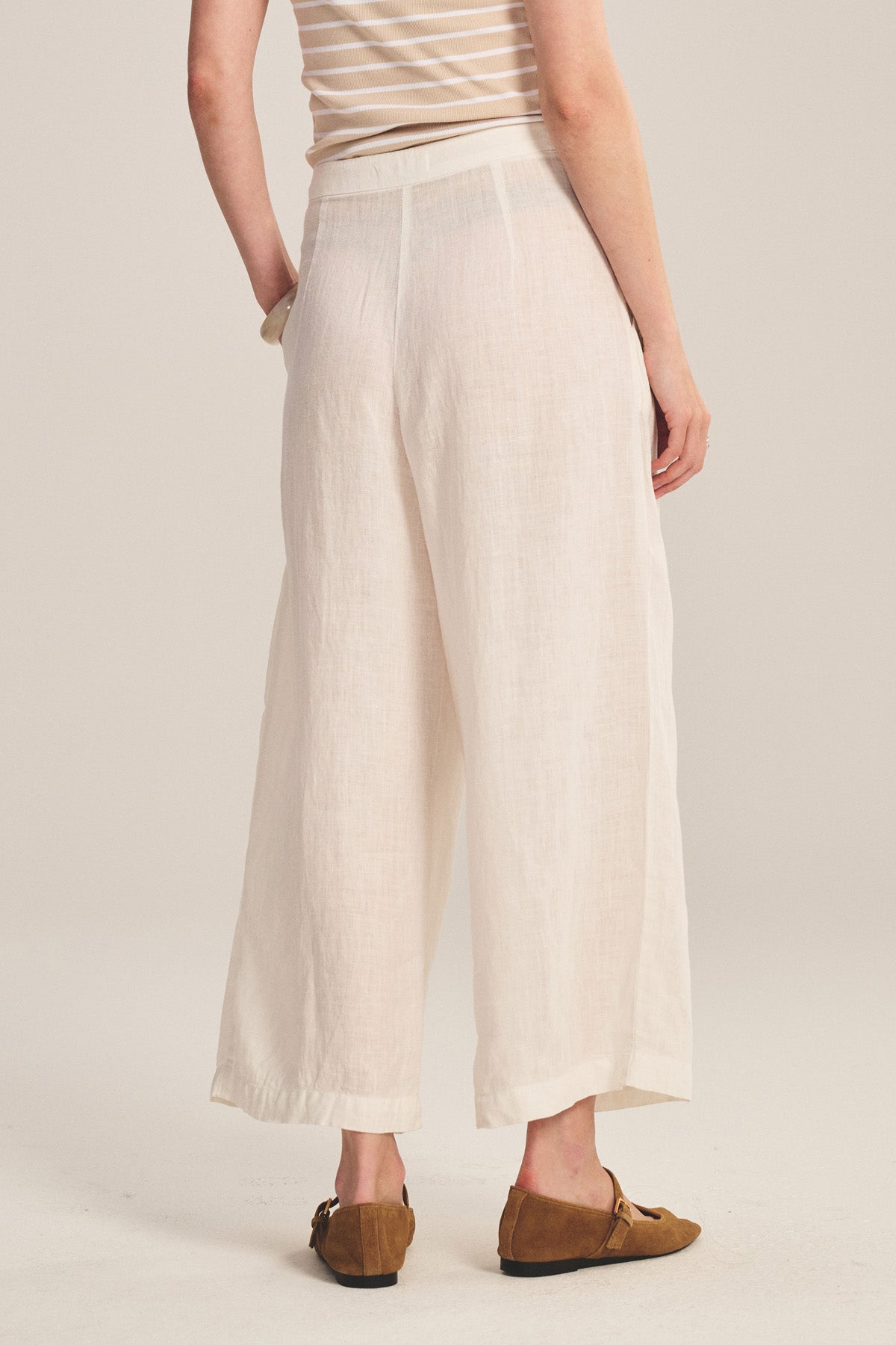 A person is seen from the back wearing Velvet by Graham & Spencer’s DANIKA LINEN PANT in a wide-leg, light color, paired with brown loafers against a neutral background.