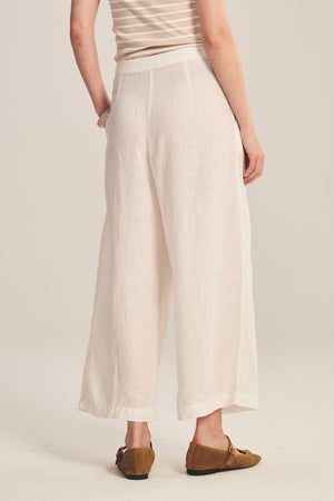 A person is seen from the back wearing Velvet by Graham & Spencer’s DANIKA LINEN PANT in a wide-leg, light color, paired with brown loafers against a neutral background.