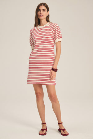 A woman wears the DARCIE DRESS by Velvet by Graham & Spencer—a vintage-inspired red and white striped terry cotton mini dress with cream trim—paired with red sandals and a red bracelet against a plain background.