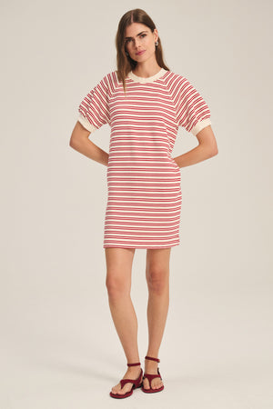 A woman stands against a plain light background wearing the Velvet by Graham & Spencer DARCIE DRESS, a vintage-inspired, short-sleeved red and white striped dress, styled with red sandals.
