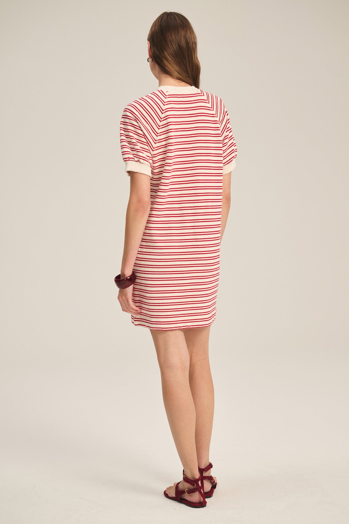 A person faces away, wearing the Velvet by Graham & Spencer DARCIE DRESS—vintage-inspired with red and white stripes—paired with red sandals, light brown hair, and a red bracelet.