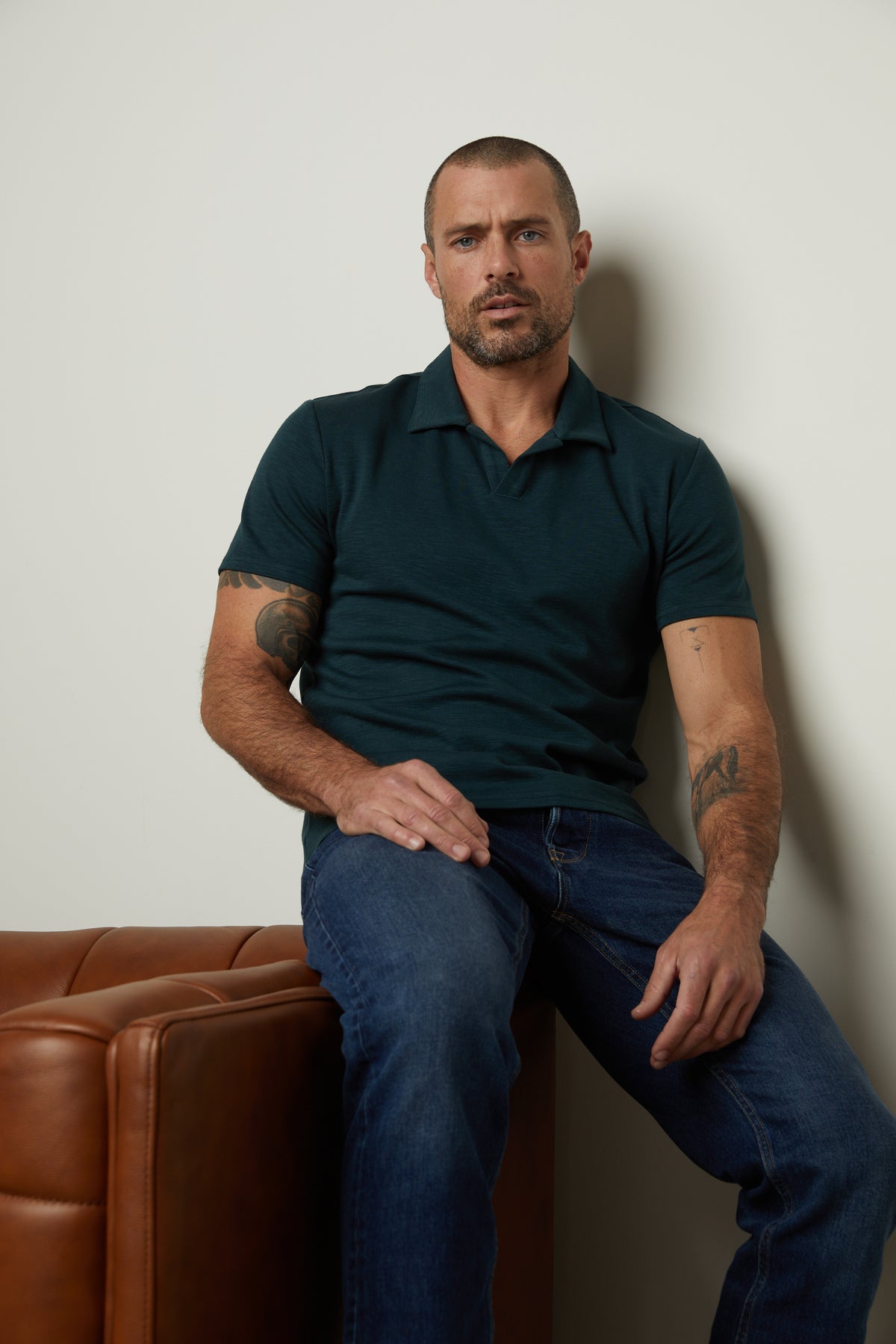   A man in jeans and a DILAN COTTON BLEND POLO by Velvet by Graham & Spencer sitting on a leather couch. 