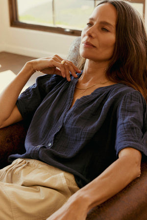 A woman with long hair is seated in a chair, wearing a dark blue DEANN COTTON GAUZE TOP by Velvet by Graham & Spencer and light-colored pants, with her left hand resting near her neck and a window in the background.