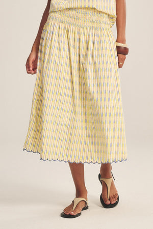A person wearing the DEBORAH SKIRT by Velvet by Graham & Spencer—a yellow and blue printed cotton skirt with a smocked elastic waist and scalloped hem—paired with black sandals.