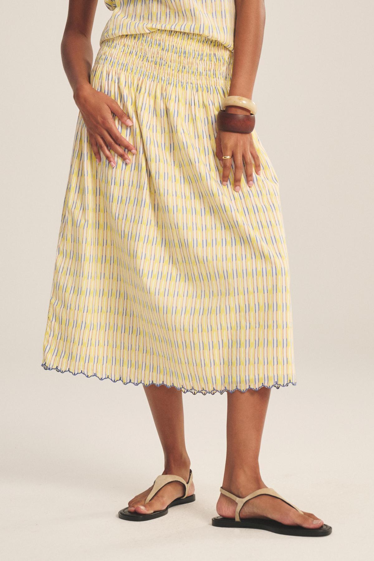 A person wearing the DEBORAH SKIRT by Velvet by Graham & Spencer—yellow and blue stripes, smocked elastic waist, scalloped hem—styled with brown bangles and beige sandals, stands against a light background.-41149936959681