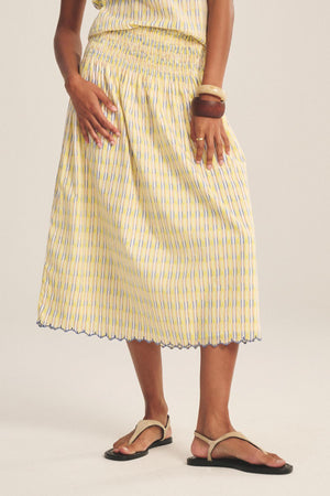 A person wearing the DEBORAH SKIRT by Velvet by Graham & Spencer—yellow and blue stripes, smocked elastic waist, scalloped hem—styled with brown bangles and beige sandals, stands against a light background.