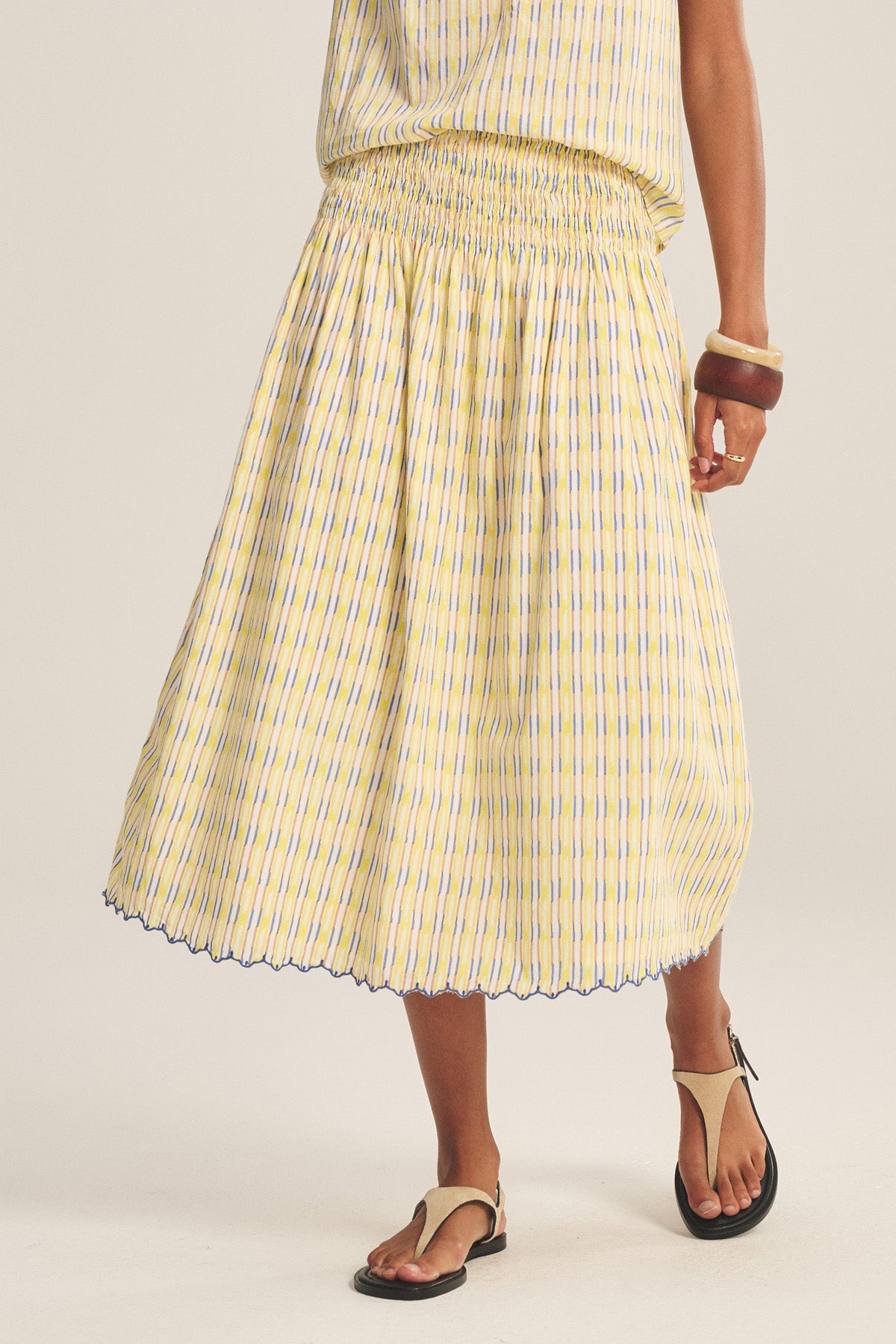 A person wears the DEBORAH SKIRT by Velvet by Graham & Spencer—a yellow and blue striped cotton skirt with a smocked elastic waist—paired with tan sandals and brown bangles, standing against a plain background.