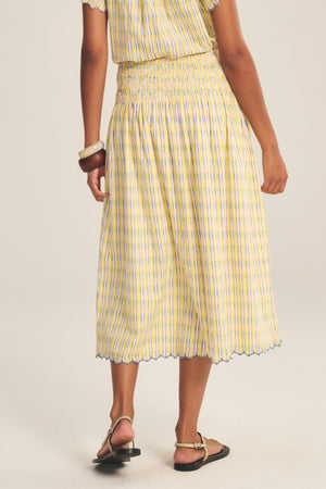 A person wears the DEBORAH SKIRT by Velvet by Graham & Spencer, a yellow and white striped cotton skirt with a scalloped hem, paired with a matching top, black sandals, and bangles on a neutral background.
