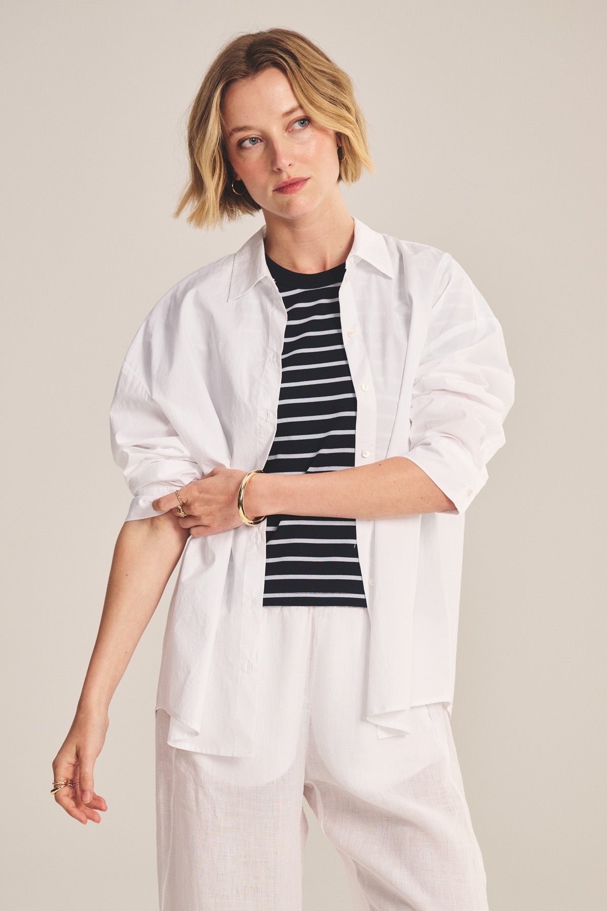 A woman wearing the DECLAN SHIRT by Velvet by Graham & Spencer, layered over a black and white striped top with white pants, stands against a neutral background and gazes to the side.-41008206282945