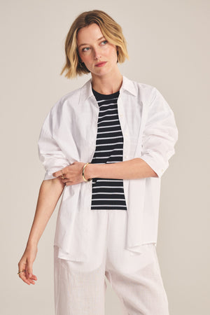 A woman wearing the DECLAN SHIRT by Velvet by Graham & Spencer, layered over a black and white striped top with white pants, stands against a neutral background and gazes to the side.