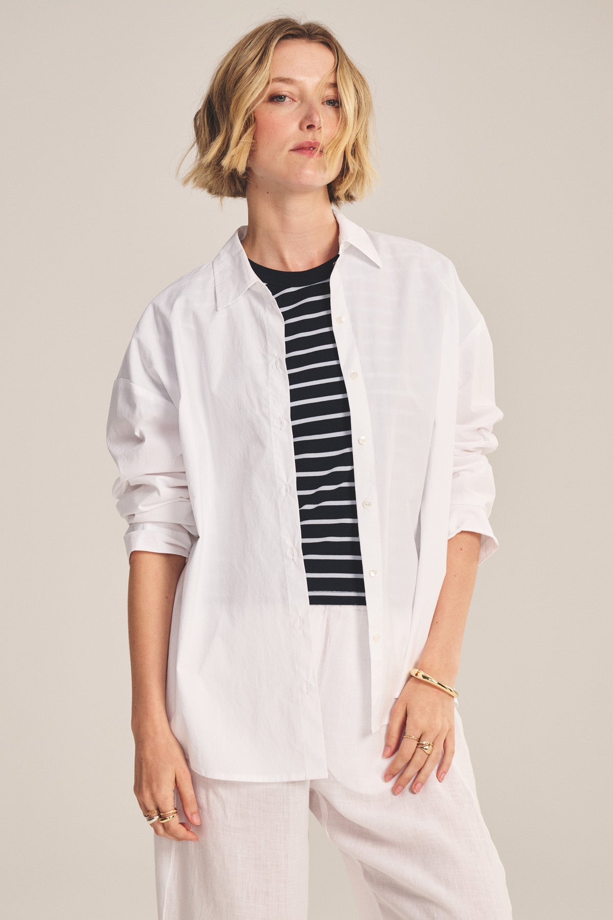 A model wears the DECLAN SHIRT by Velvet by Graham & Spencer—an oversized cotton poplin style—layered over a black and white striped top, with white pants and gold jewelry, posed against a plain background.