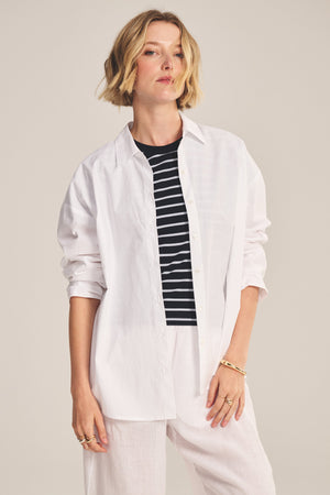 A model wears the DECLAN SHIRT by Velvet by Graham & Spencer—an oversized cotton poplin style—layered over a black and white striped top, with white pants and gold jewelry, posed against a plain background.