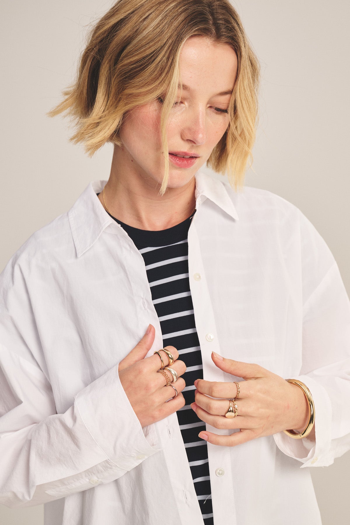 A person with short blonde hair wears the oversized white DECLAN SHIRT by Velvet by Graham & Spencer over a black and white striped top, accessorized with multiple rings and a gold bracelet.-41008206020801