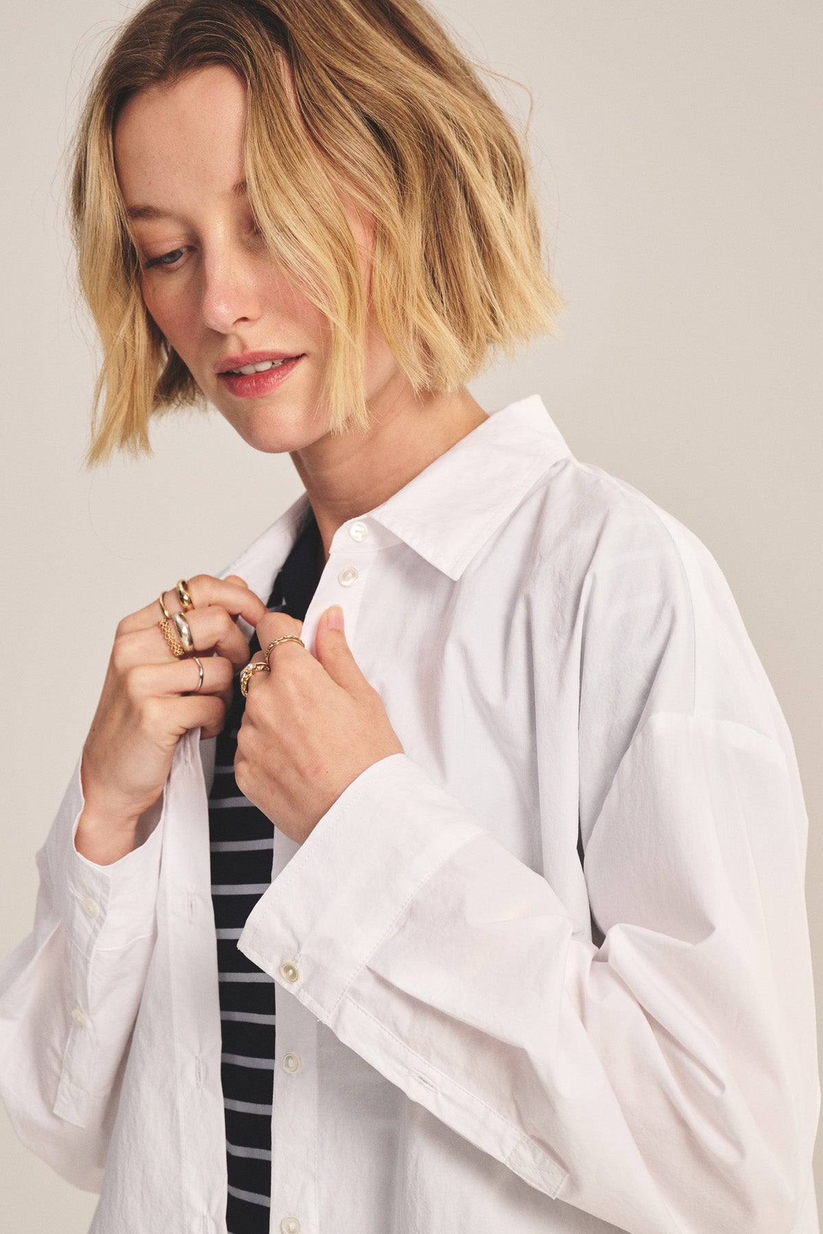 A woman with short blonde hair wears the DECLAN SHIRT by Velvet by Graham & Spencer—an oversized white cotton poplin shirt—layered over a black and white striped top, as she looks down and adjusts her collar.-41008205955265