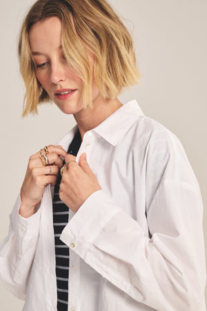 A woman with short blonde hair wears the DECLAN SHIRT by Velvet by Graham & Spencer—an oversized white cotton poplin shirt—layered over a black and white striped top, as she looks down and adjusts her collar.