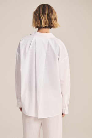A person with short, wavy hair stands facing away against a plain background, wearing the DECLAN SHIRT by Velvet by Graham & Spencer—an oversized cotton poplin shirt—paired with light-colored pants.