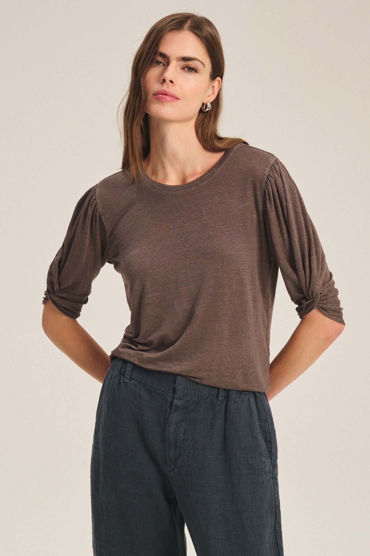 A woman stands in a neutral pose wearing the DEE TEE by Velvet by Graham & Spencer, featuring ruched sleeves and dark, loose linen pants, set against a plain background.