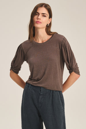 A woman stands in a neutral pose wearing the DEE TEE by Velvet by Graham & Spencer, featuring ruched sleeves and dark, loose linen pants, set against a plain background.
