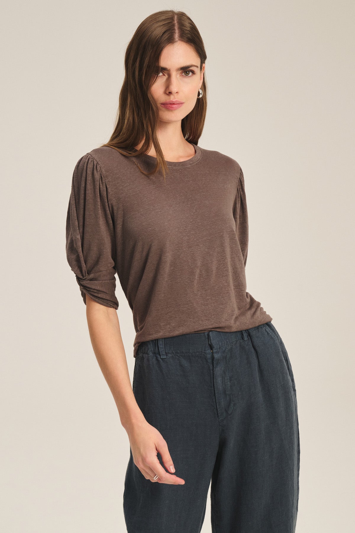 A woman wears the DEE TEE by Velvet by Graham & Spencer, featuring ruched sleeves and crafted from a linen-elastane blend, paired with dark loose pants as she stands against a plain background.