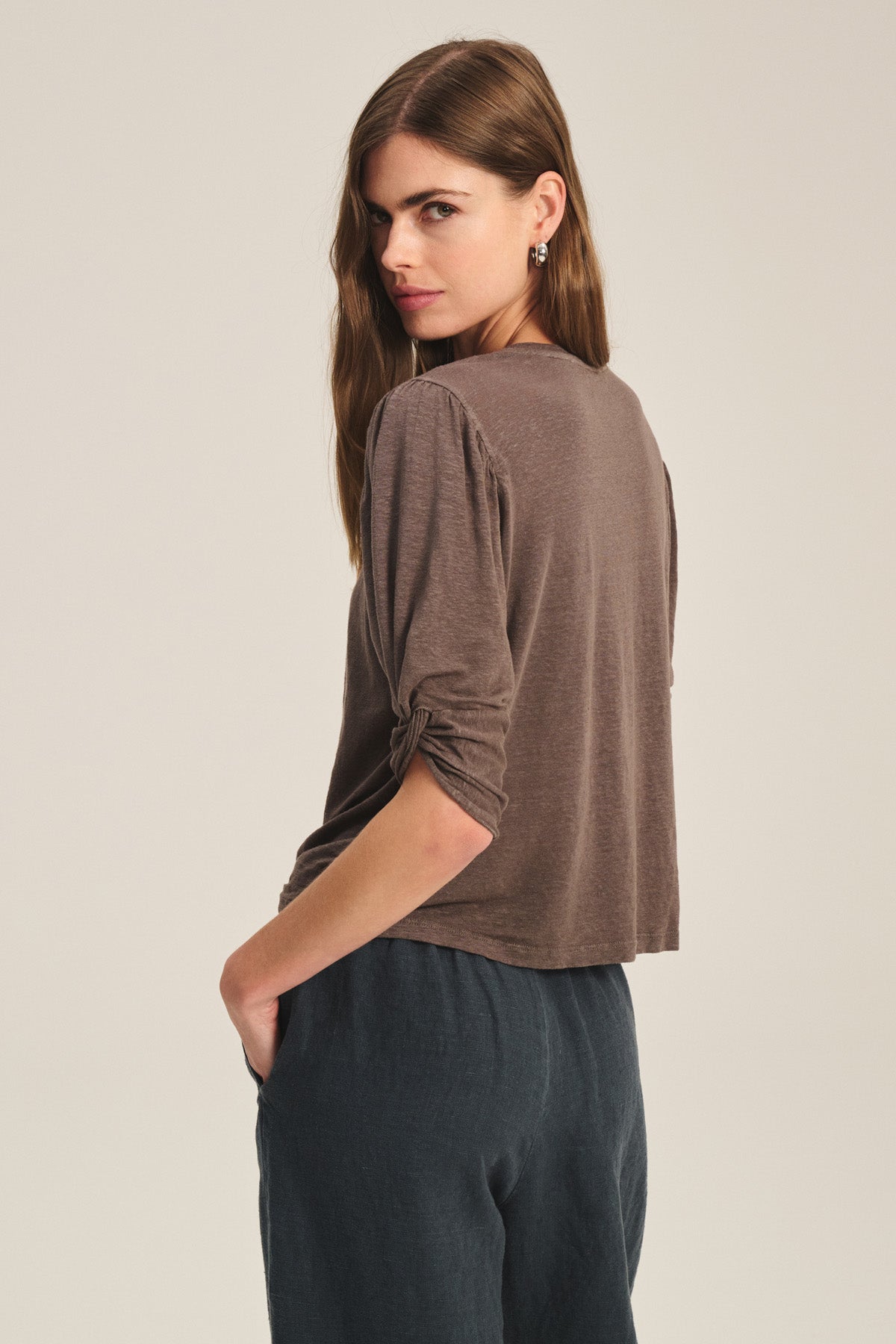 A woman with long brown hair stands sideways, wearing the DEE TEE by Velvet by Graham & Spencer—a brown linen-elastane blend tee with ruched sleeves—and dark pants, looking over her shoulder.