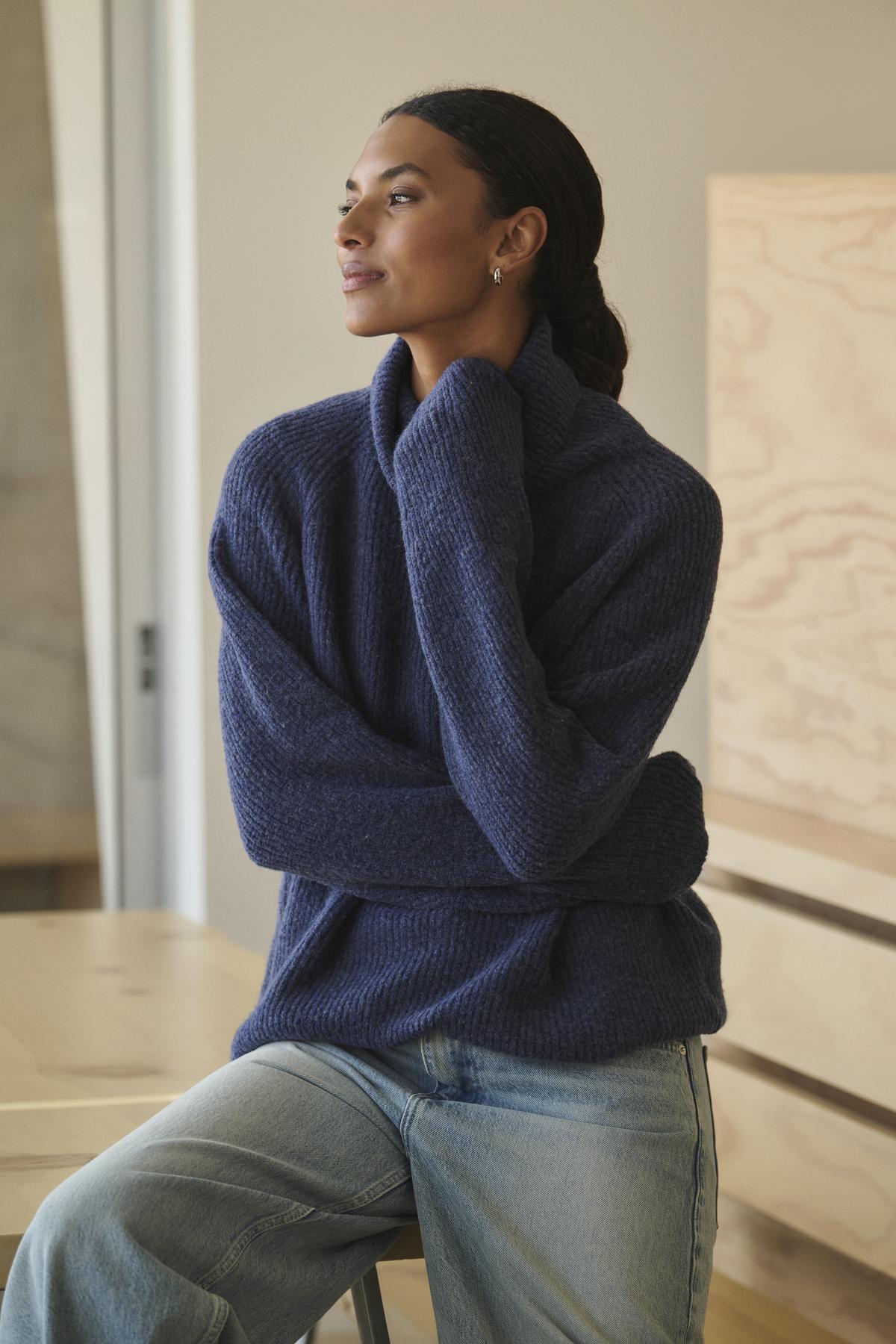   Wearing a cozy JUDITH TURTLENECK SWEATER by Velvet by Graham & Spencer along with jeans, a person sits on a stool, glancing to the side with a neutral expression. 