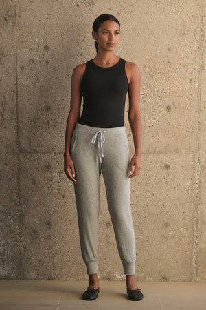 A person stands against a concrete wall wearing a black tank top and the KOKO RIBBED JOGGERS in gray, featuring a white drawstring waist by Velvet by Graham & Spencer, complemented by black ballet flats.