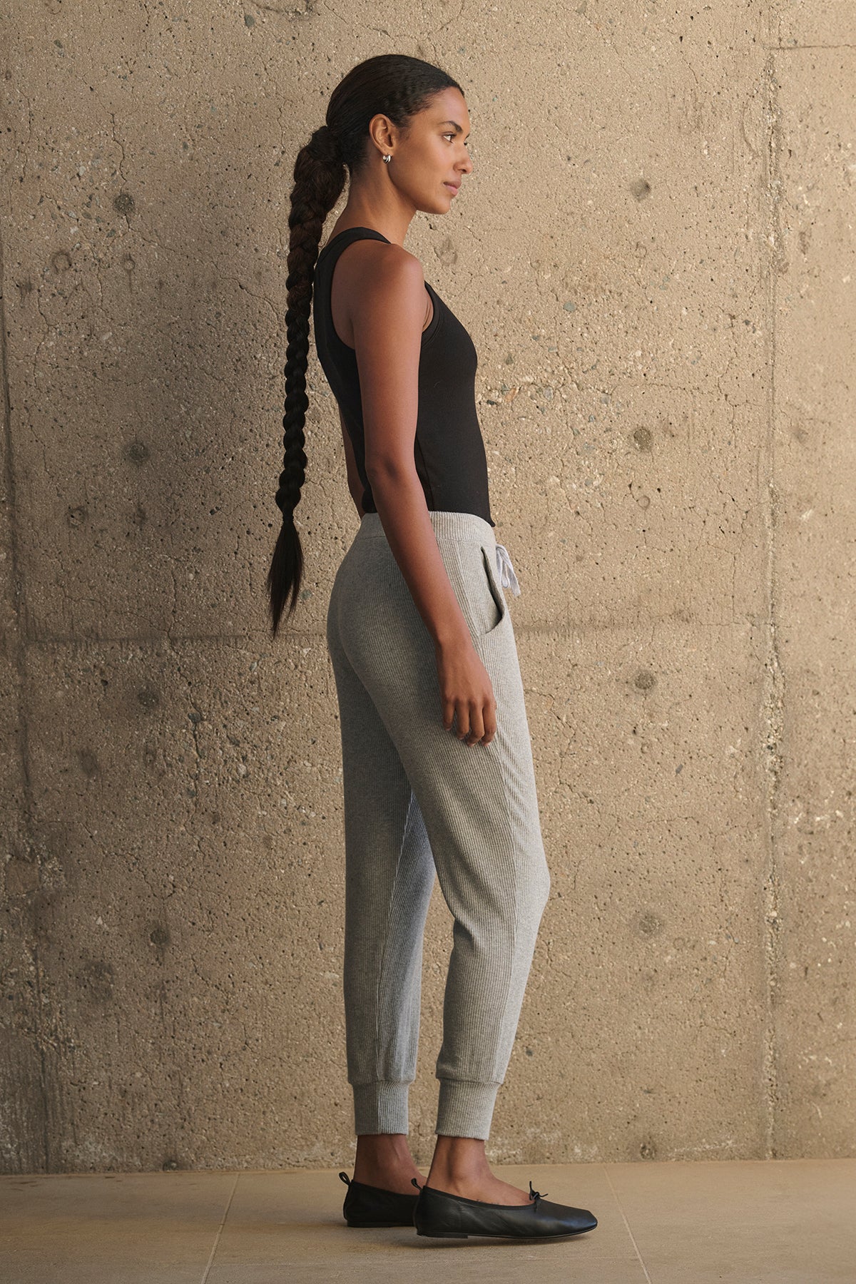   A person with a long braid wearing a black tank top, KOKO RIBBED JOGGERS by Velvet by Graham & Spencer with a drawstring waist, and black shoes stands in profile against a textured concrete wall. 