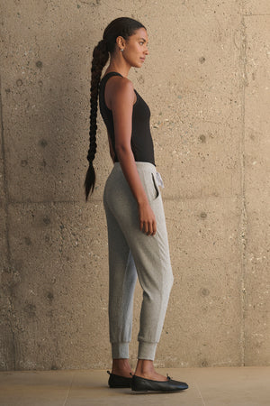 A person with a long braid wearing a black tank top, KOKO RIBBED JOGGERS by Velvet by Graham & Spencer with a drawstring waist, and black shoes stands in profile against a textured concrete wall.