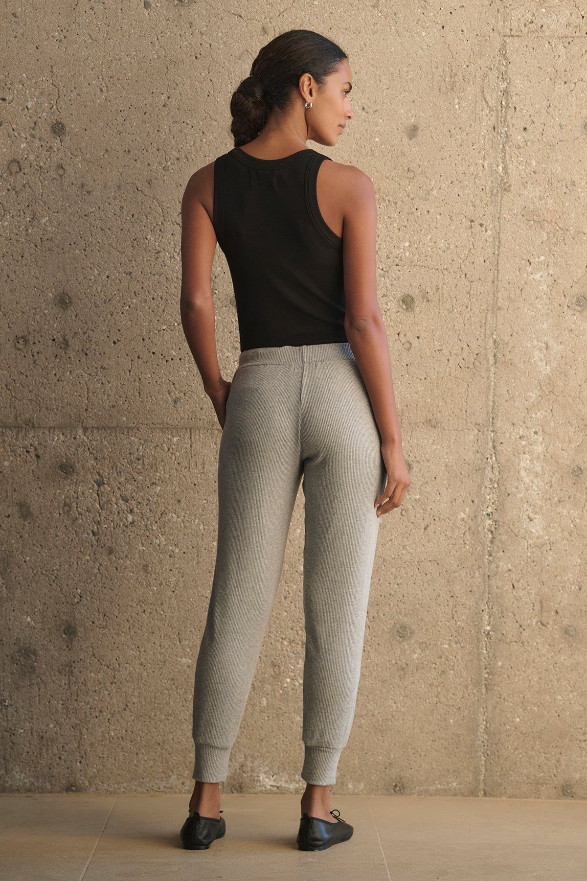 A person stands facing a textured wall, wearing a black tank top and the KOKO RIBBED JOGGERS by Velvet by Graham & Spencer, complemented by black shoes.-38282007871681