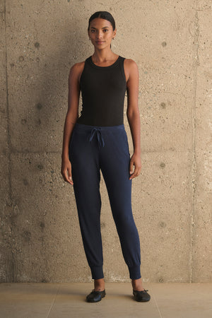 A person stands against a concrete wall, wearing a black sleeveless top, KOKO RIBBED JOGGERS in navy by Velvet by Graham & Spencer, and black shoes.