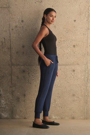 A woman with a long braid stands against a textured concrete wall, wearing a black tank top, the KOKO RIBBED JOGGERS from Velvet by Graham & Spencer featuring a drawstring waist in blue, and black loafers.