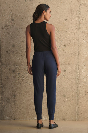 A person wearing a black tank top and Velvet by Graham & Spencer's KOKO RIBBED JOGGERS in navy, featuring a drawstring waist, stands with their back to the camera against a textured beige wall.