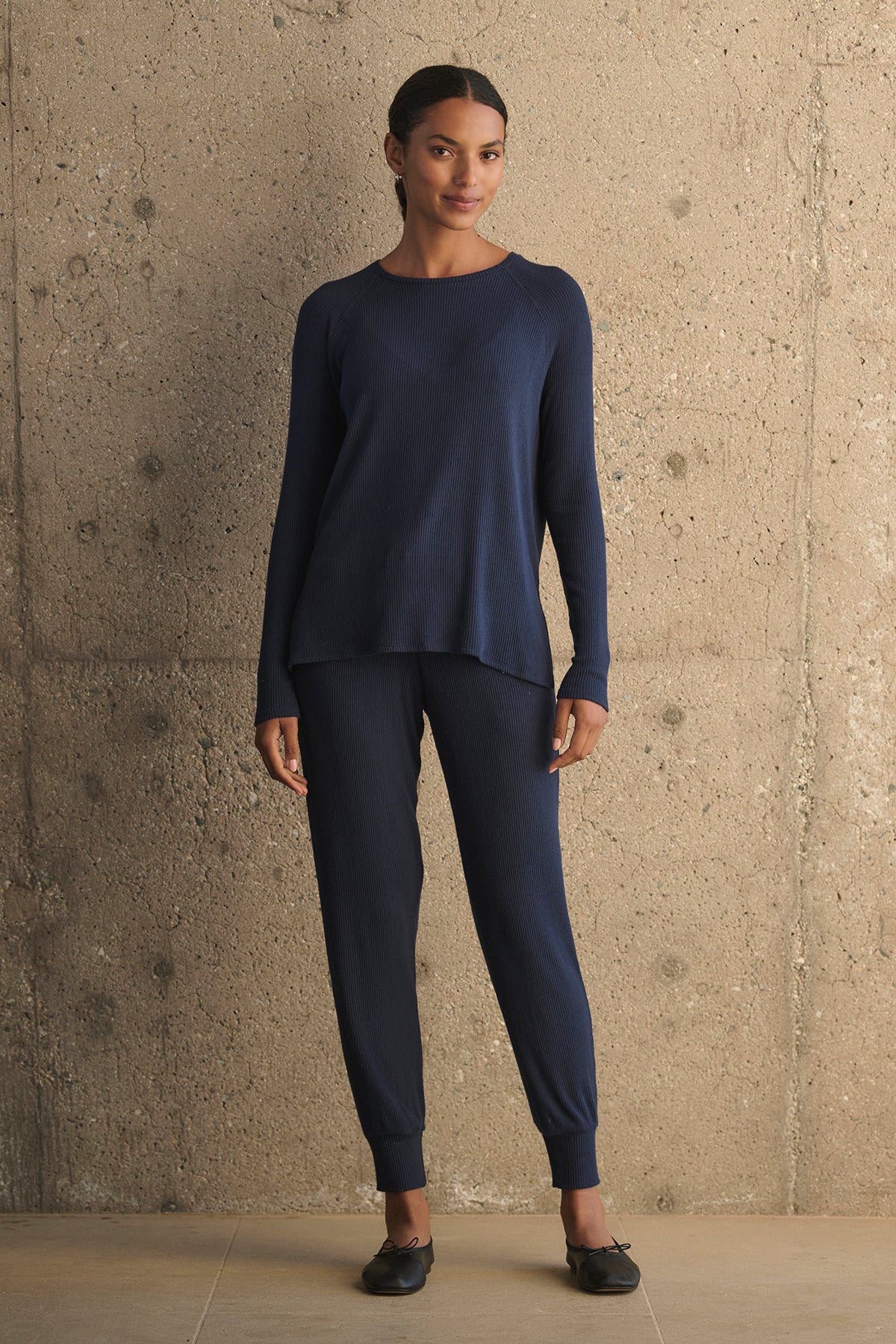   A person wearing a matching navy blue sweater and KOKO RIBBED JOGGERS with a drawstring waist from Velvet by Graham & Spencer stands against a textured concrete wall. 