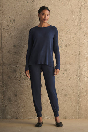 A person wearing a matching navy blue sweater and KOKO RIBBED JOGGERS with a drawstring waist from Velvet by Graham & Spencer stands against a textured concrete wall.