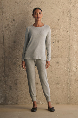 A person dressed in Velvet by Graham & Spencer's KOKO Ribbed Joggers and a coordinating gray long-sleeve top stands against a textured concrete wall, with their hands at their sides and black shoes on their feet.